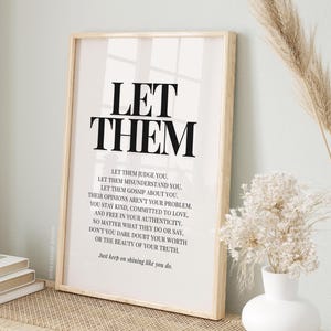 以下が含まれることがあります： 太字の黒い文字で「LET THEM」と書かれた額入りのプリント。その下に、「Let them judge you... Just keep on shining like you do」と書かれた小さなテキストがあります。プリントは、装飾的な要素のある織物の表面に表示されています。