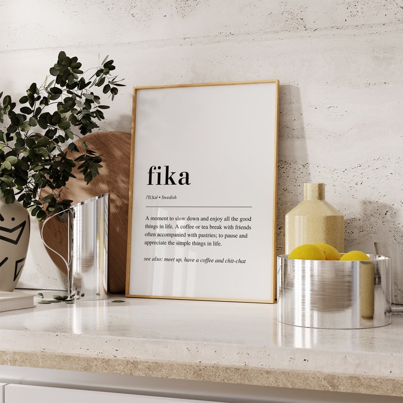 May include: A framed print with a gold frame and white background. The print features the word "fika" in black text with a definition in smaller text below. The definition explains that "fika" is a Swedish word for a coffee or tea break with friends, often accompanied by pastries.