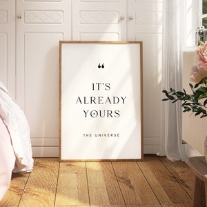 May include: A framed print with a white background and black text that reads "It's already yours. The Universe." The print is hanging on a white wall in a room with a pink blanket and a vase of pink flowers.