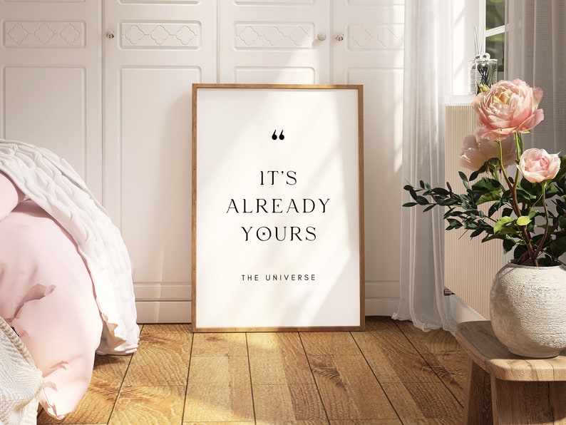 May include: A framed print with a white background and black text that reads "It's already yours. The Universe." The print is hanging on a white wall in a room with a pink blanket and a vase of pink flowers.