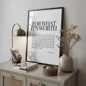 Può includere: Una stampa incorniciata con il testo "FOR WHAT IT'S WORTH". Una lampada da scrivania in stile vintage, riviste, un piccolo cestino intrecciato e un vaso in ceramica con fiori secchi sono su un mobile grigio.
