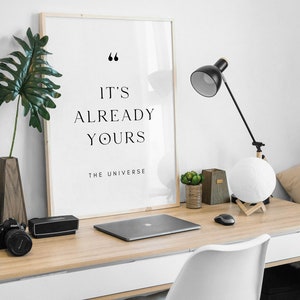 May include: A framed print with the text "It's already yours - The Universe" in a minimalist style. The print is on a desk with a laptop, a lamp, and a potted plant.