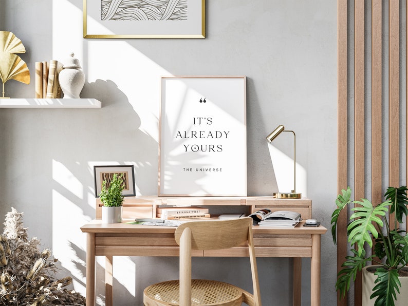May include: A framed print with the text "It's already yours - The Universe" on a white background. The print is sitting on a wooden desk with a gold lamp, a potted plant, and a few books.