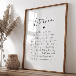 Peut inclure: Une affiche encadrée avec l'inscription "Let them" en écriture cursive au-dessus d'un cœur. En dessous, un texte inspirant est écrit dans une police simple. Le cadre est marron chaud, et le fond est blanc.