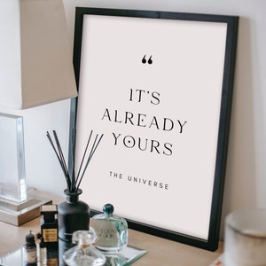 May include: A black framed print with a white background and the text "It's already yours - The Universe" in black font.