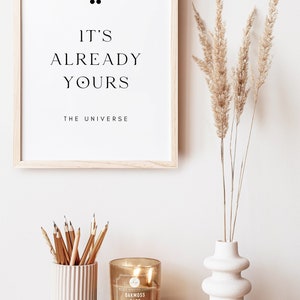 May include: A framed print with the text "It's already yours, The Universe."  A vase with dried pampas grass, a candle, and a pencil holder are on a wooden table.