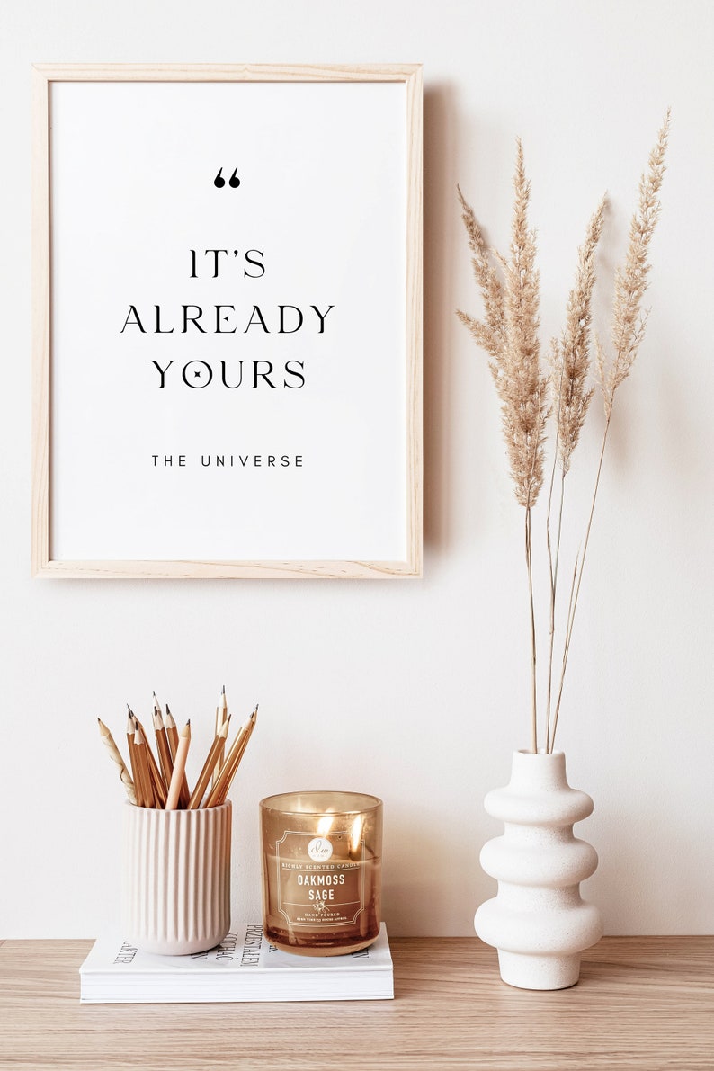 May include: A framed print with the text "It's already yours, The Universe."  A vase with dried pampas grass, a candle, and a pencil holder are on a wooden table.