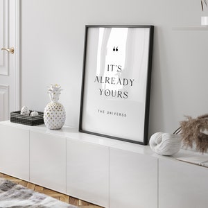 May include: A black framed print with the text "It's already yours - The Universe" on a white background. The print is displayed on a white cabinet with a white pineapple and a white seashell.