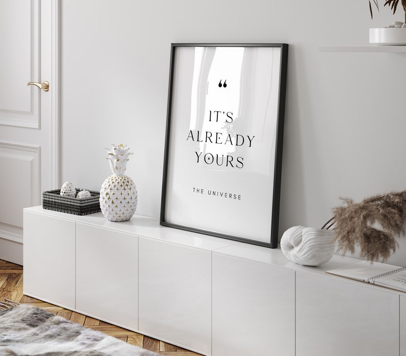 May include: A black framed print with the text "It's already yours - The Universe" on a white background. The print is displayed on a white cabinet with a white pineapple and a white seashell.