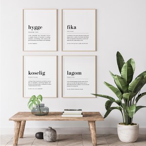 May include: Four black and white framed prints with the words "hygge", "fika", "koselig", and "lagom" defined. The prints are hanging on a white wall above a wooden table with a potted plant.