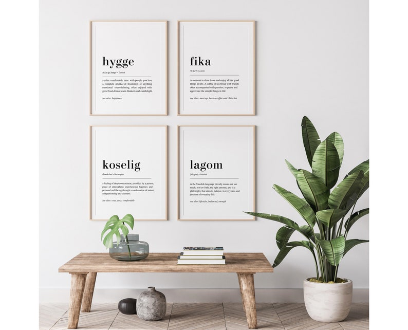 May include: Four black and white framed prints with the words "hygge", "fika", "koselig", and "lagom" defined. The prints are hanging on a white wall above a wooden table with a potted plant.