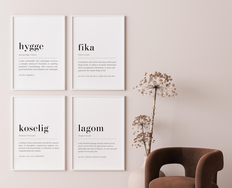 May include: Four framed prints with white mounts and black frames on a pale pink wall. The prints feature the words "hygge", "fika", "koselig", and "lagom" with their definitions in English. The prints are arranged in a 2x2 grid.