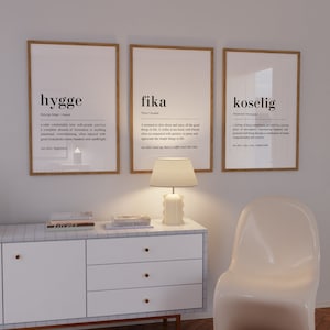 May include: Three framed prints with white backgrounds and black text. The prints define the words "hygge", "fika", and "koselig" and their meanings. The prints are hanging on a white wall above a white dresser with three drawers and a white chair.