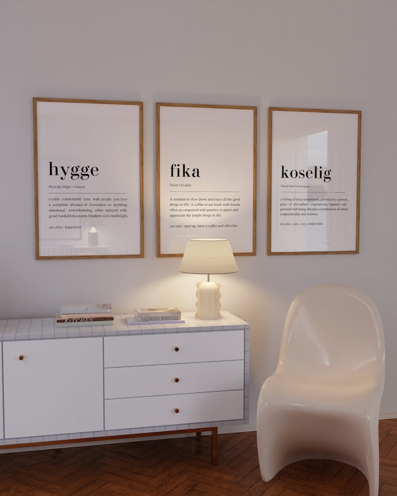 May include: Three framed prints with white backgrounds and black text. The prints define the words "hygge", "fika", and "koselig" and their meanings. The prints are hanging on a white wall above a white dresser with three drawers and a white chair.