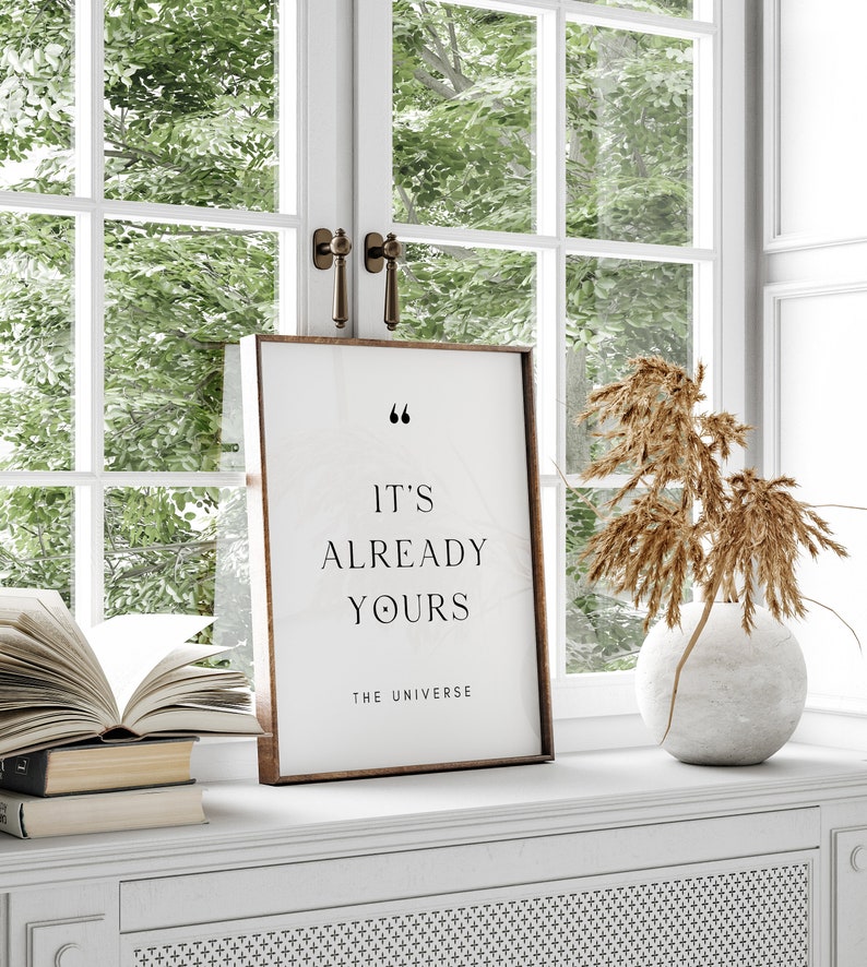 May include: A framed print with a white background and black text that reads "It's already yours - The Universe". The print is sitting on a white shelf with a book and a vase with dried flowers.