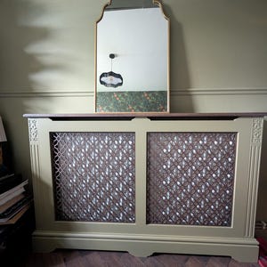 May include: A sage green radiator cover with a decorative lattice design. A gold-framed mirror with an arched top is mounted above the cover. The mirror reflects a patterned wallpaper and a black pendant light.