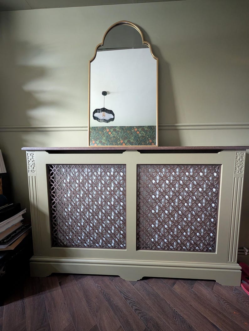 May include: A sage green radiator cover with a decorative lattice design. A gold-framed mirror with an arched top is mounted above the cover. The mirror reflects a patterned wallpaper and a black pendant light.