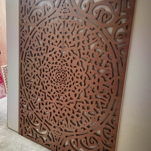 May include: Two-panel decorative wooden screen with intricate, symmetrical cut-out patterns. The screen is a warm brown color, with a complex floral or mandala-like design. The panels are leaning against a white wall, suggesting an interior design element.