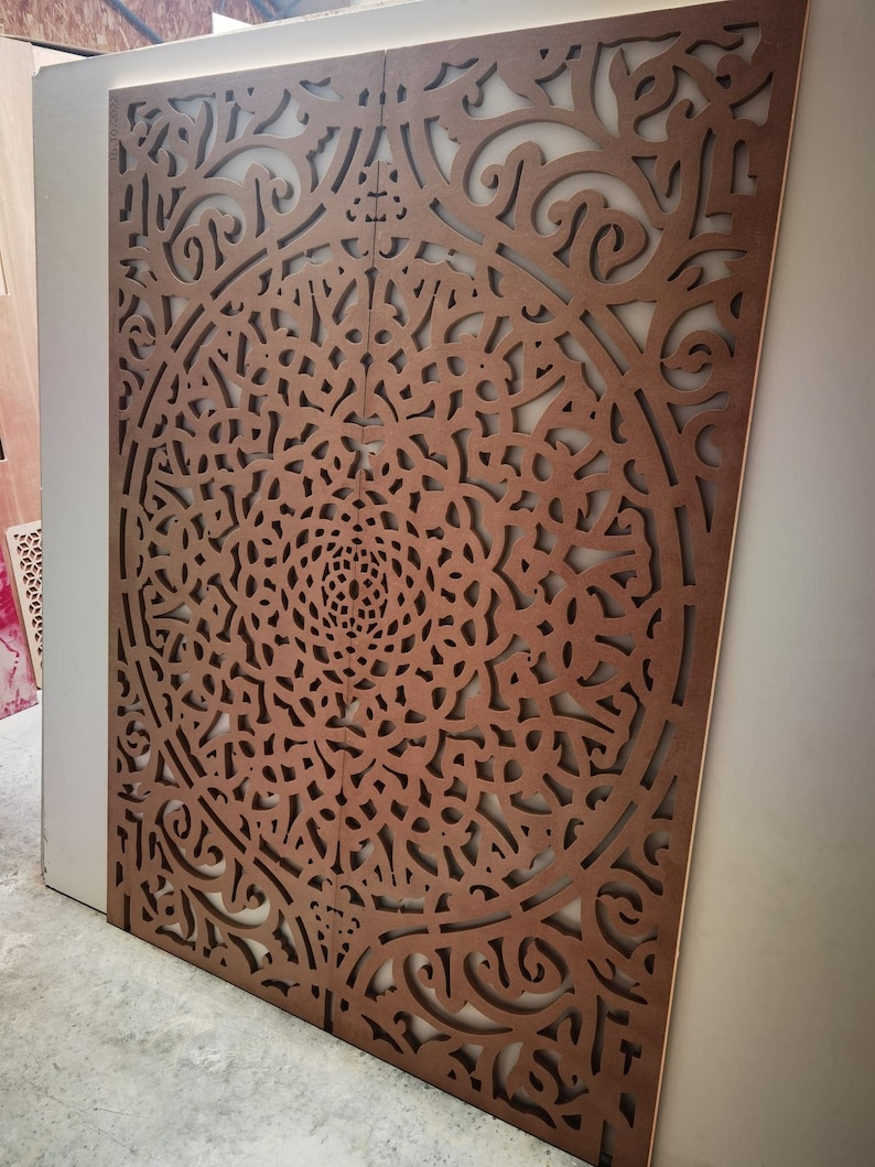 May include: Two-panel decorative wooden screen with intricate, symmetrical cut-out patterns. The screen is a warm brown color, with a complex floral or mandala-like design. The panels are leaning against a white wall, suggesting an interior design element.