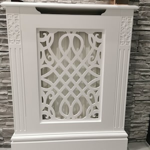 May include: White wooden radiator cover with a decorative, intricate, cut-out design. The cover has a raised base and a flat top.