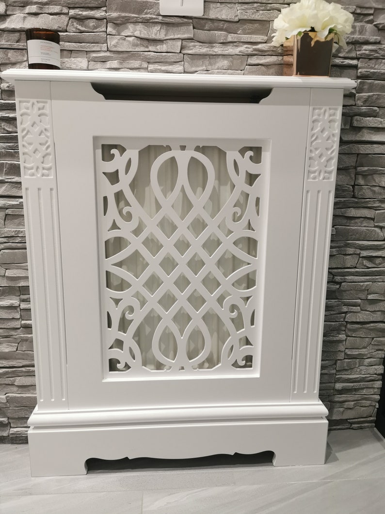 May include: White wooden radiator cover with a decorative, intricate, cut-out design. The cover has a raised base and a flat top.