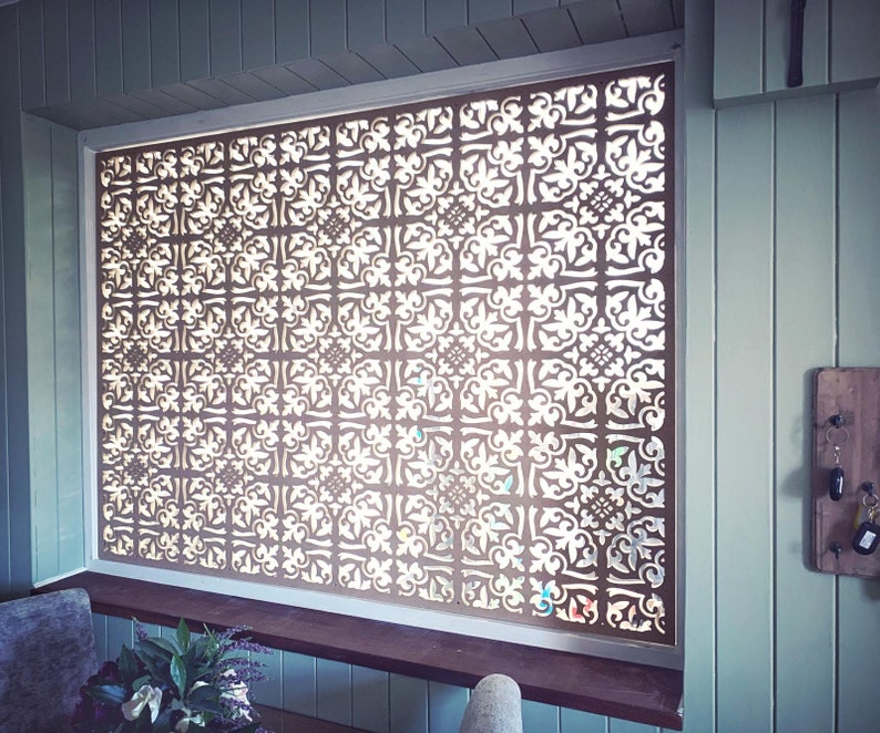 May include: A decorative window screen with a repeating, ornate pattern. The screen is made of a dark material and is set within a white frame. The intricate design allows light to filter through, creating a visually appealing effect.