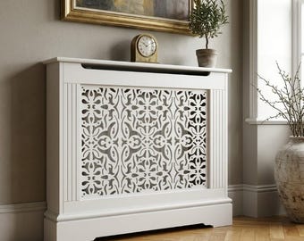 May include: A white radiator cover with a decorative, openwork floral design. The cover features a flat top, vertical side panels, and a base. A small clock and potted plant are on top. This is a decorative home decor item, often used to conceal radiators.