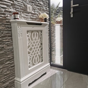 May include: White radiator cover with a decorative, intricate design. The cover is freestanding and has a base with a curved front edge. The cover is in front of a stone wall.
