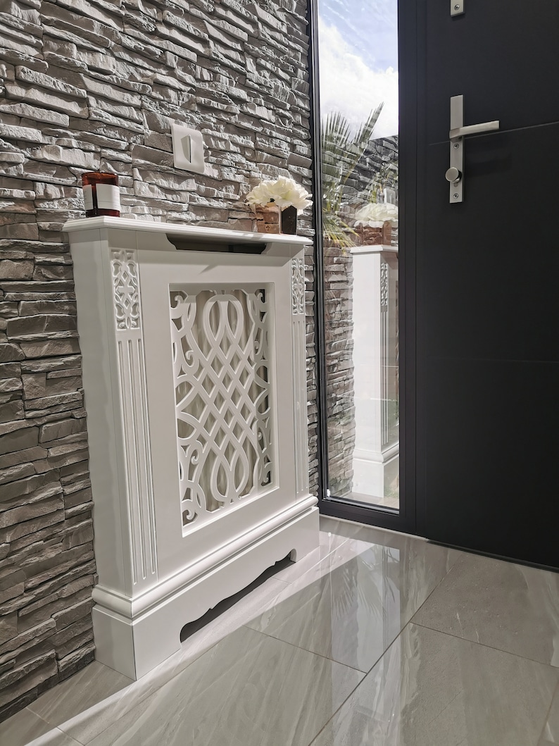 May include: White radiator cover with a decorative, intricate design. The cover is freestanding and has a base with a curved front edge. The cover is in front of a stone wall.