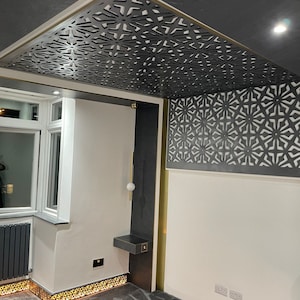 May include: Interior shot featuring a decorative ceiling with a geometric pattern in dark gray. The ceiling design is complemented by gold trim and a matching wall panel. The room has white walls, a gray carpet, and a modern light fixture.