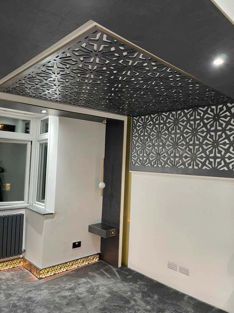 May include: Interior shot featuring a decorative ceiling with a geometric pattern in dark gray. The ceiling design is complemented by gold trim and a matching wall panel. The room has white walls, a gray carpet, and a modern light fixture.