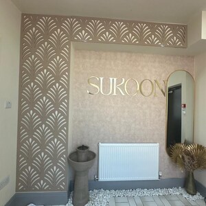 May include: Interior design featuring a wall with a decorative leaf pattern in beige and white. The word "SUKOON" is displayed in gold lettering. A mirror, radiator, and decorative vase are also visible.