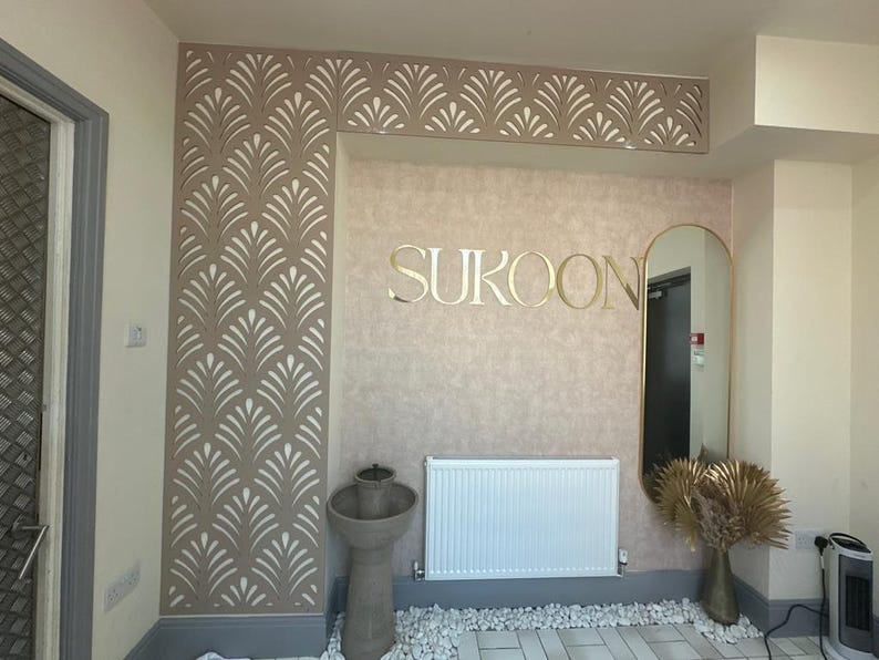 May include: Interior design featuring a wall with a decorative leaf pattern in beige and white. The word "SUKOON" is displayed in gold lettering. A mirror, radiator, and decorative vase are also visible.