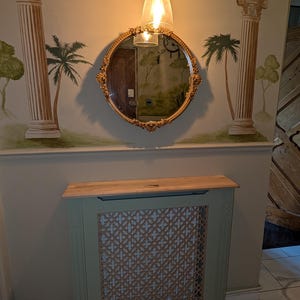 May include: A wall-mounted mirror with a gold frame, a light fixture, and a radiator cover. The mirror is round with decorative edges. The radiator cover is painted sage green with a lattice pattern. The wall has painted columns and palm trees.