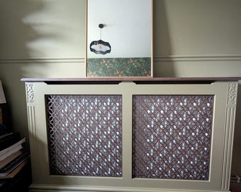 May include: A sage green radiator cover with a decorative lattice design. A gold-framed mirror with an arched top is mounted above the cover. The mirror reflects a patterned wallpaper and a black pendant light.