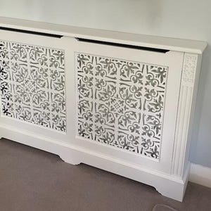 May include: White radiator cover with a decorative, patterned front. The cover has a rectangular shape with a flat top and a base with a slight curve. The front panels feature a repeating floral design.
