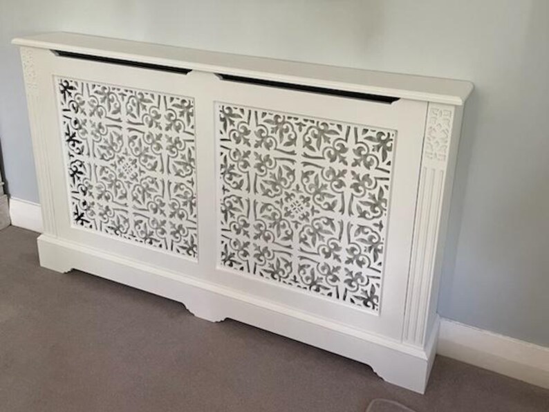 May include: White radiator cover with a decorative, patterned front. The cover has a rectangular shape with a flat top and a base with a slight curve. The front panels feature a repeating floral design.