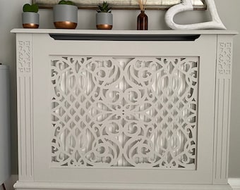 May include: White painted radiator cover with a decorative, intricate, cut-out design. The cover has a base with a scalloped edge. Three small potted plants and a white heart-shaped ornament are on top of the cover.