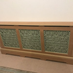 May include: A wooden radiator cover with three panels featuring a decorative cut-out design. The panels are painted a light green color and the cover is a light brown color.