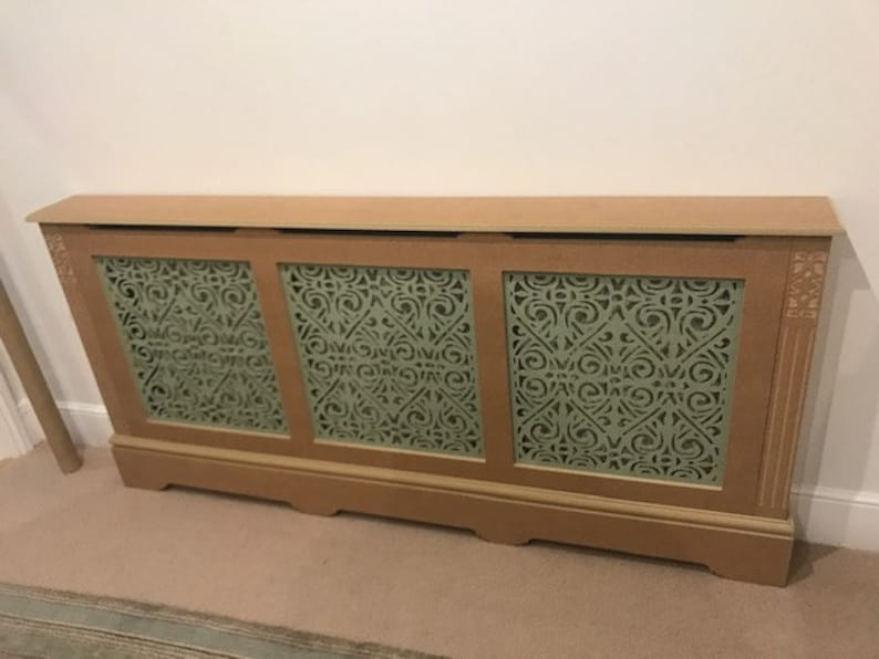 May include: A wooden radiator cover with three panels featuring a decorative cut-out design. The panels are painted a light green color and the cover is a light brown color.