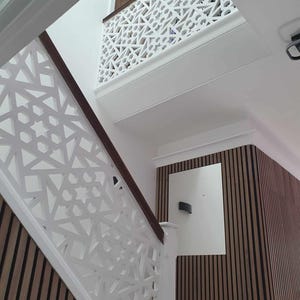 May include: Interior shot of a staircase with white geometric patterned railings and a dark wood handrail. The wall features vertical wood paneling. The ceiling has recessed lighting.
