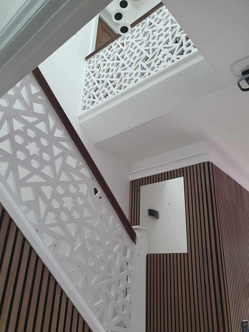 May include: Interior shot of a staircase with white geometric patterned railings and a dark wood handrail. The wall features vertical wood paneling. The ceiling has recessed lighting.