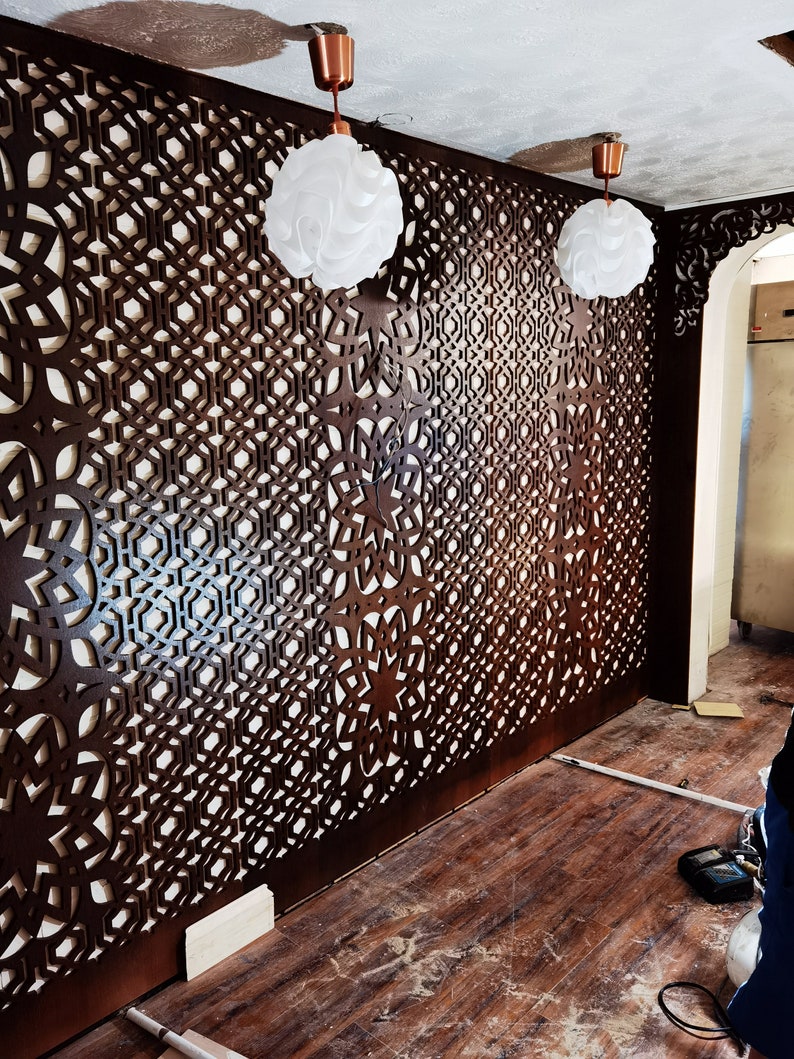May include: A room with dark brown, intricately patterned wooden wall panels. Two white, spherical pendant lights hang from the ceiling. The floor is made of wooden planks. The design is geometric and creates a decorative screen.