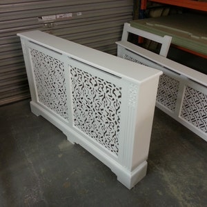 May include: Two white wooden radiator covers with intricate cut-out designs. The covers are freestanding and have a decorative top.