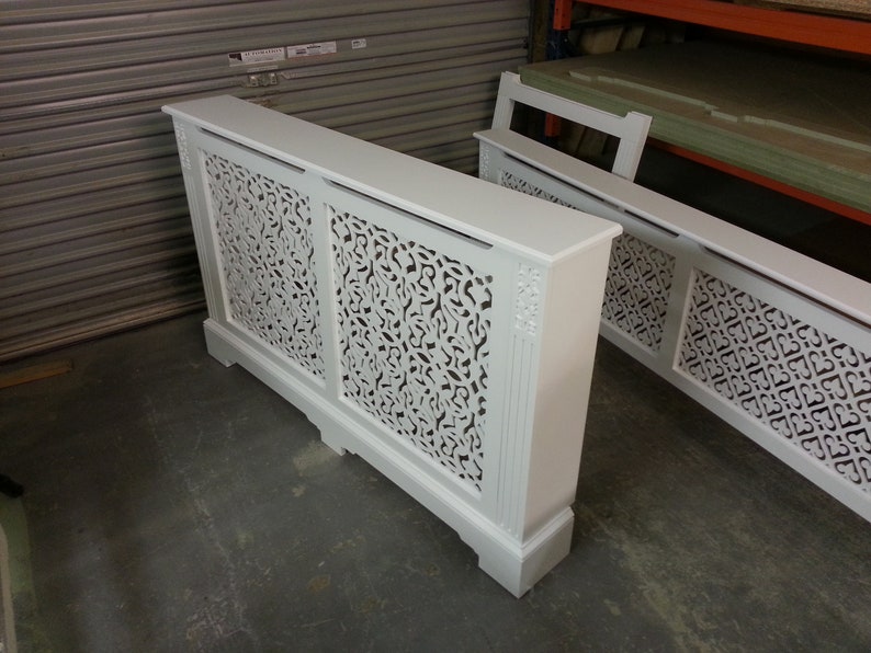May include: Two white wooden radiator covers with intricate cut-out designs. The covers are freestanding and have a decorative top.