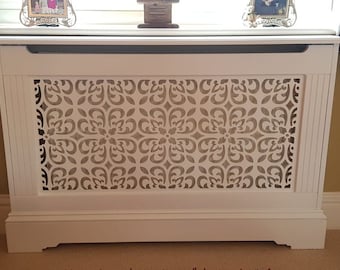 May include: White radiator cover with a decorative, cut-out design. The cover has a raised top and a base with a decorative edge.