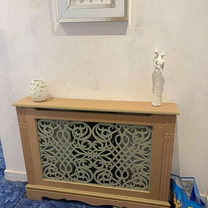 May include: A beige radiator cover with a decorative green lattice design. The cover has a wooden frame and sits on a blue carpet. A white ceramic figurine of two people holding hands is on top of the cover.