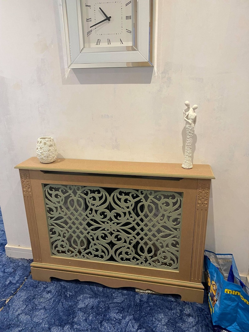 May include: A beige radiator cover with a decorative green lattice design. The cover has a wooden frame and sits on a blue carpet. A white ceramic figurine of two people holding hands is on top of the cover.