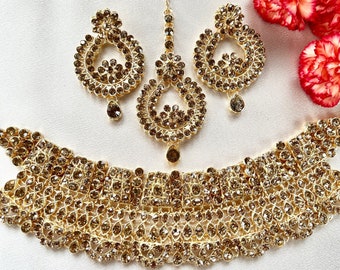 Pode incluir: Um conjunto de joias douradas com colar, brincos e um adorno de cabeça. O conjunto é adornado com inúmeras pedras pequenas e brilhantes. Os brincos e o adorno de cabeça têm um pendente em forma de gota. As joias são exibidas em uma superfície branca.