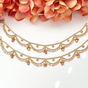 Puede incluir: Tobilleras doradas con cadenas delicadas, pequeños detalles de perlas y abalorios ornamentados. Las tobilleras se exhiben sobre un fondo blanco con flores rosas y melocotón en la parte superior. Una hermosa pieza de joyería.